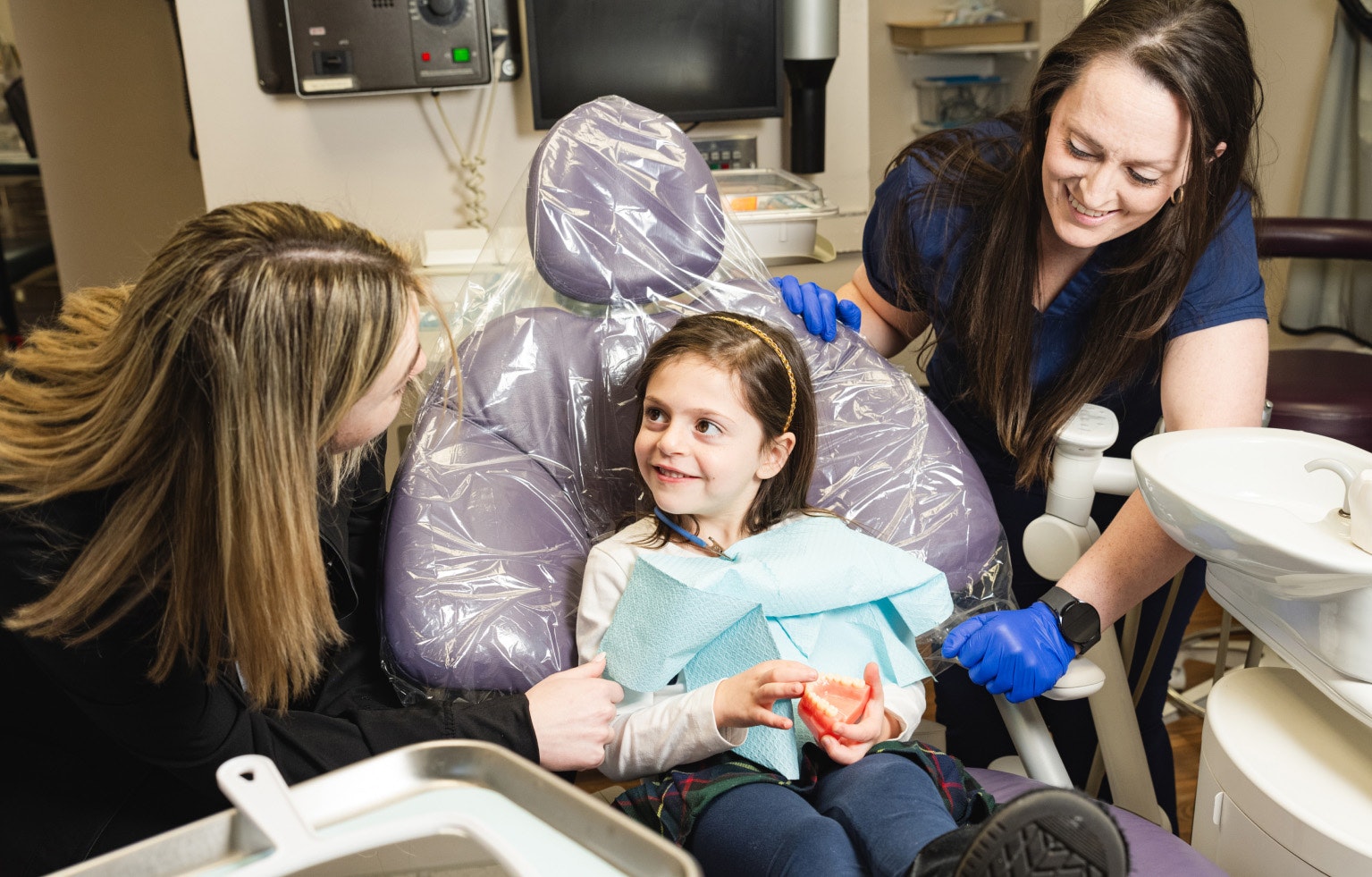A child in a dental chair being attended to by two dental assistants
